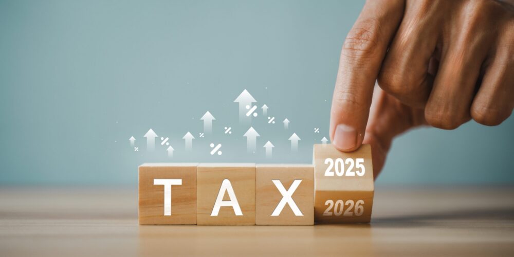 Hand placing a block labeled 2025 above wooden blocks spelling TAX and 2026, representing tax planning and future tax strategy.